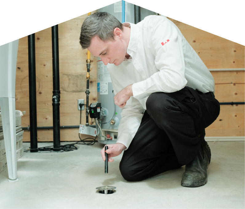 Mr. Furnace plumbing specialist inspecting a basement floor drain with a flashlight during a drain cleaning service, in a utility room with visible pipes, water heater, and wood panel walls. Mr. Furnace plumbing specialist inspecting a basement floor drain with a flashlight during a drain cleaning service, in a utility room with visible pipes, water heater, and wood panel walls.