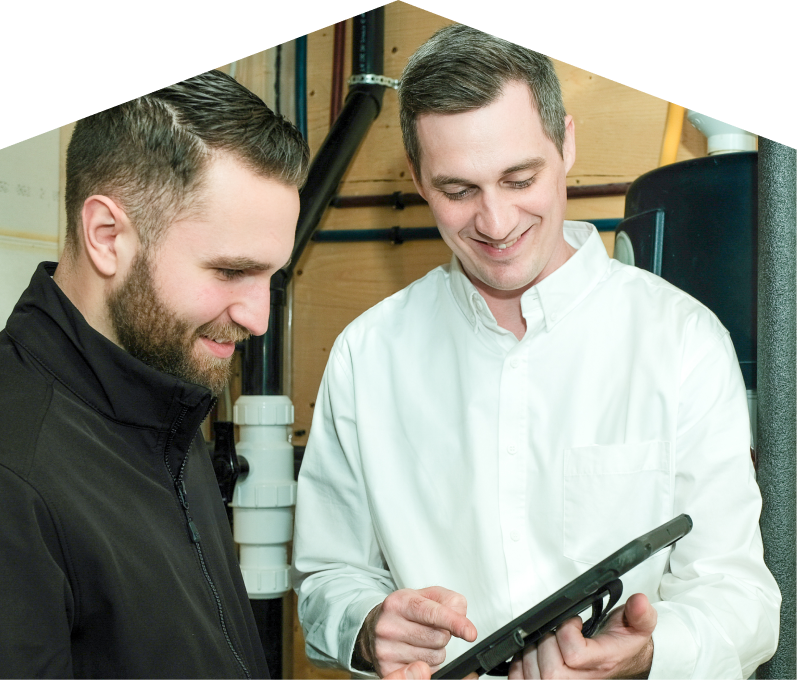 Mr. Furnace specialist reviewing maintenance plan options with a smiling homeowner using a tablet, inside a utility room with visible piping. Mr. Furnace specialist reviewing maintenance plan options with a smiling homeowner using a tablet, inside a utility room with visible piping.
