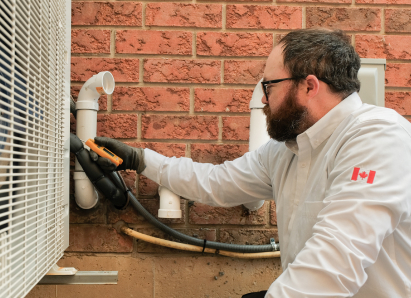 A Mr. Furnace HVAC specialist wearing glasses and gloves uses a diagnostic tool to service an outdoor AC unit beside a brick wall. A Mr. Furnace HVAC specialist wearing glasses and gloves uses a diagnostic tool to service an outdoor AC unit beside a brick wall.
